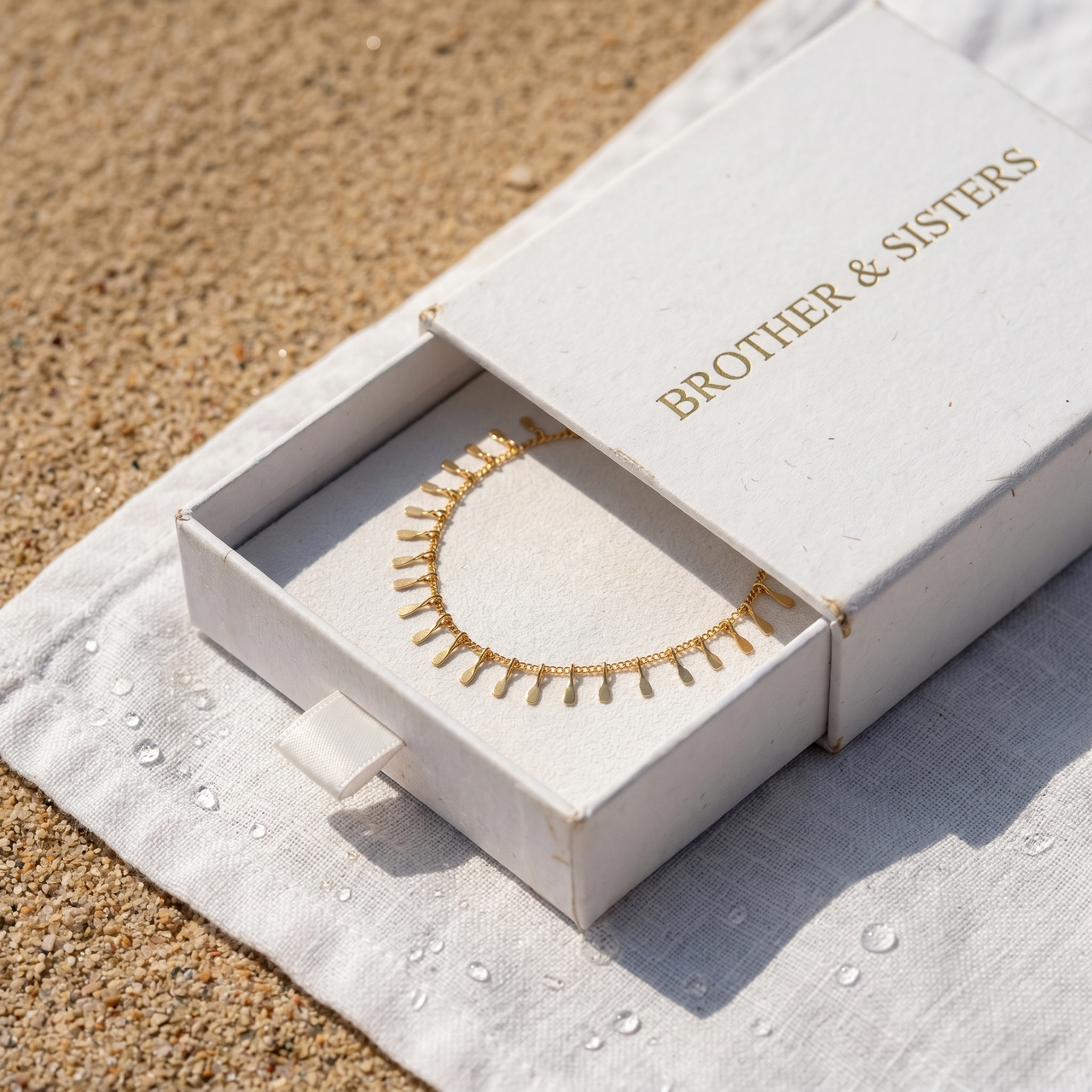 Sun Ray Fringe Anklet in a Brother & Sisters box on sandy background, showcasing delicate gold design and summer vibes.