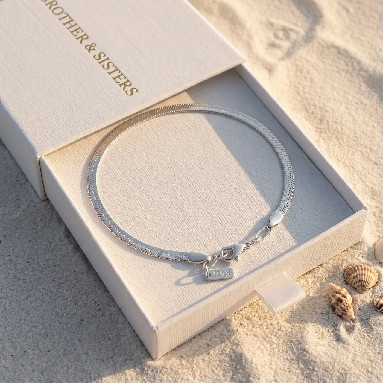 Layla silver anklet inside B&S signature white slide-drawer gift box on beach sand at golden hour with sea shell — flat herringbone chain closed oval, fermoir and B&S tag visible, luxury silver anklet packaging