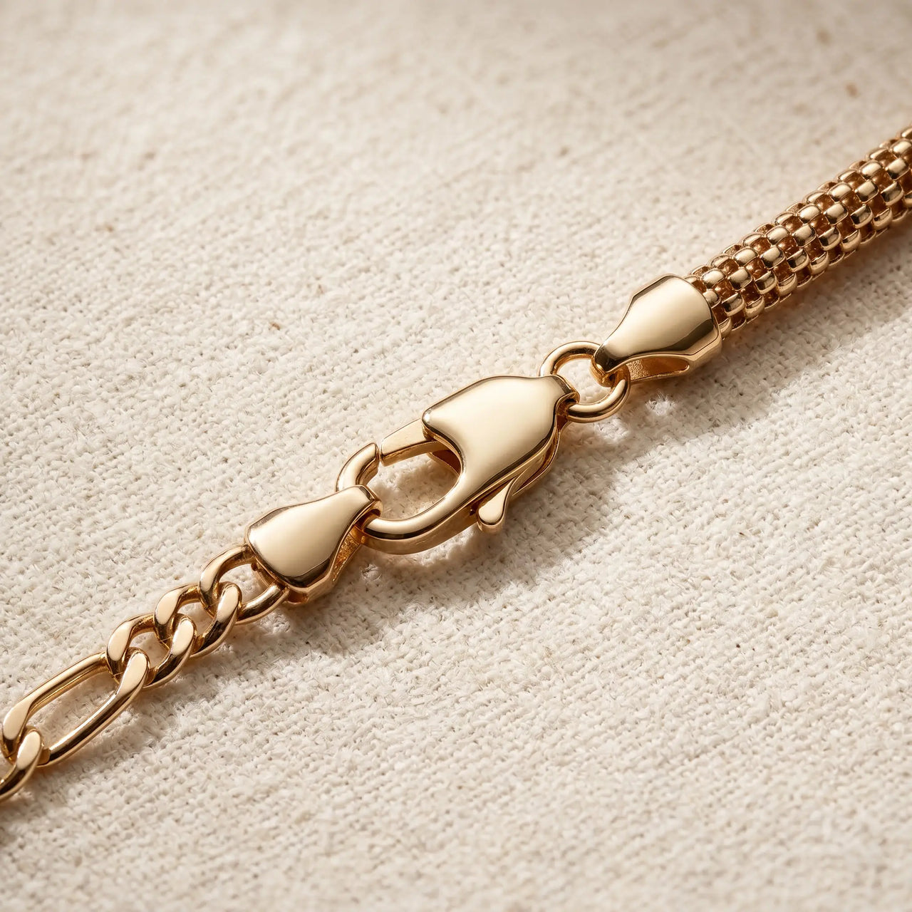 Maëlle gold anklet macro detail — swivel lobster clasp + figaro extension chain + tube-mesh popcorn weave on cream linen, premium 18K gold PVD hardware