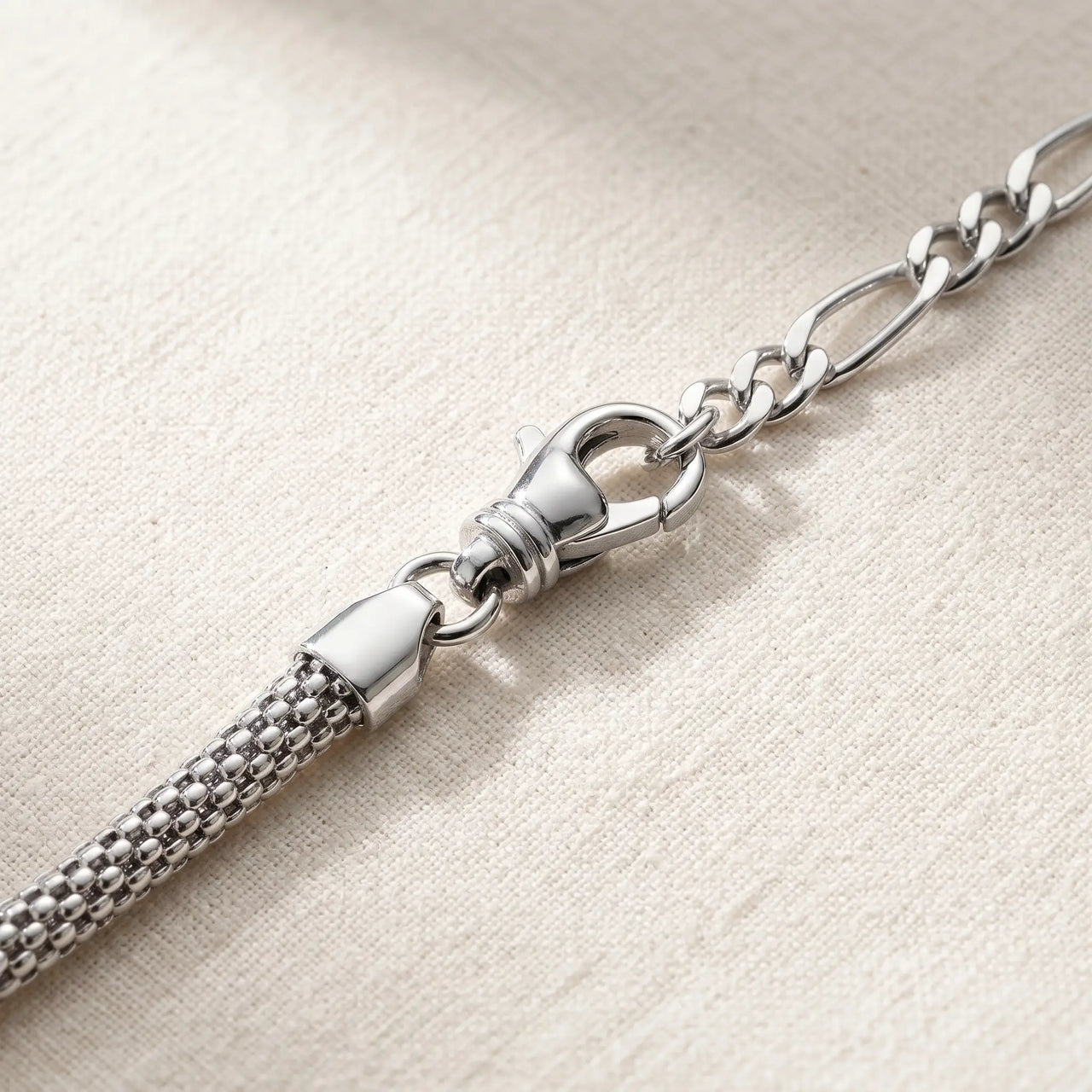 Maëlle silver anklet macro detail — polished swivel lobster clasp + figaro extension chain + tube-mesh popcorn woven cylindrical chain on cream linen, premium hardware