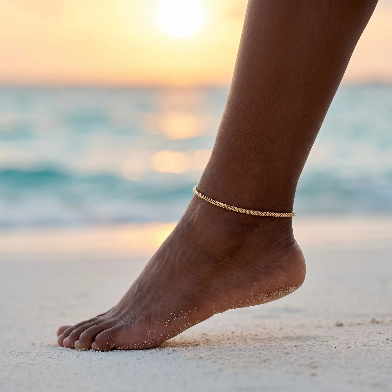 Maëlle delicate gold tube-mesh anklet on Caribbean Black model ankle at Maldives sunset — golden hour beach, refined feminine proportions